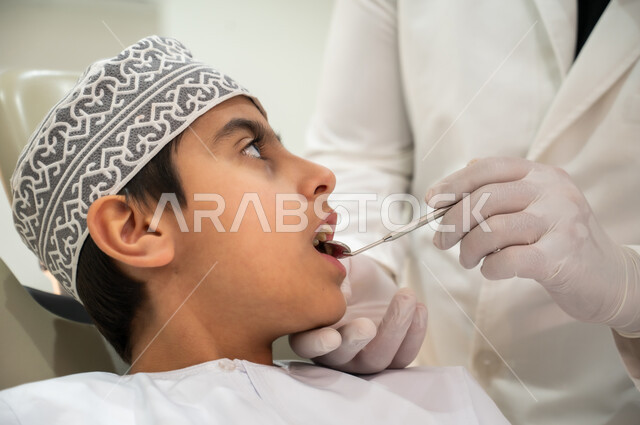 المعدات والأدوات المعقمة، الزيارة الدورية للعيادات الصحية، المحافظة على الصحة الفموية، طبيب أسنان يرتدي المعطف والقفازات الطبية يقوم بفحص فم المريض، صورة مقربة من الجانب لولد عربي خليجي عماني يرتدي الكمه يجلس في العيادة السنية يتلقى العلاج اللازم
