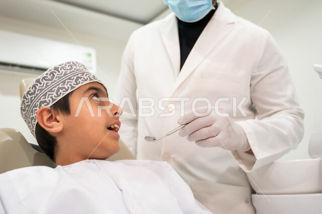 المحافظة على الصحة الفموية، الأدوات والمعدات المعقمة، زيارة العيادات الصحية لاتخاذ الإجراءات اللازمة، طبيب أسنان يرتدي المعطف والقفازات والكمامة الطبية يمسك بيده أداة الفحص، صورة مقربة لولد عربي خليجي عماني يرتدي الكمه يجلس في العيادة السنية يتلقى العلاج اللازم