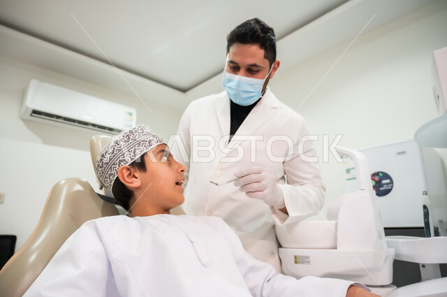 زيارة العيادات الصحية لاتخاذ الإجراءات اللازمة، المحافظة على الصحة الفموية، الأدوات والمعدات المعقمة، طبيب أسنان يرتدي المعطف والقفازات والكمامة الطبية يمسك بيده أداة الفحص، صورة مقربة لولد عربي خليجي عماني يرتدي الكمه يجلس في العيادة السنية يتلقى العلاج اللازم