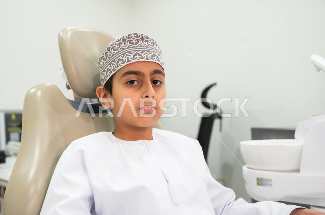 زيارة العيادات الصحية لاتخاذ الإجراءات اللازمة، المحافظة على الصحة الفموية، النظر إلى الكاميرا بتعابير الجدية والثقة بالنفس، صورة مقربة لولد عربي خليجي عماني يرتدي الكمه يجلس في العيادة السنية يتلقى العلاج اللازم