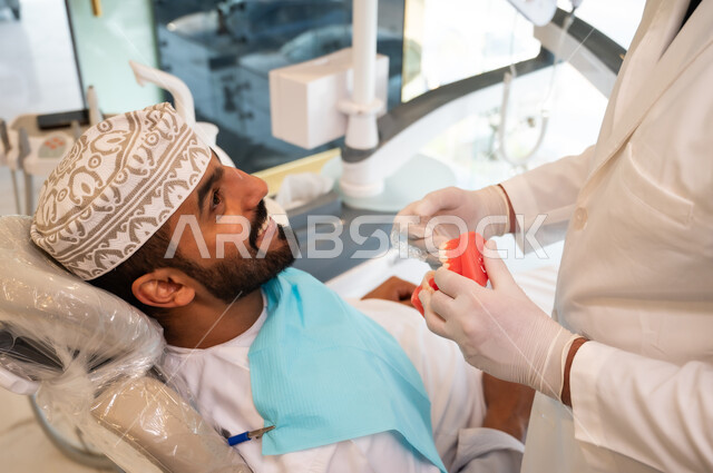 Explaining the treatment plan, a dentist wearing a coat and medical gloves holding a dental model, expressions of pleasure and reassurance, providing health care in specialized centers, a top view of an Arab Gulf Omani man sitting in the dental clinic receiving the necessary treatment for his mouth