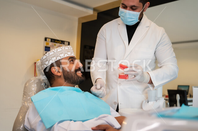 تعابير السرور والاطمئنان، تقديم الرعاية الصحية في المراكز التخصصية، طبيب أسنان يرتدي المعطف والكمامة الطبية يمسك بيده نموذج سني، رجل عربي خليجي عماني يجلس في العيادة السنية يتلقى العلاج اللازم للحصول على ابتسامة مثالية، إعطاء الإرشادات والتعليمات اللازمة