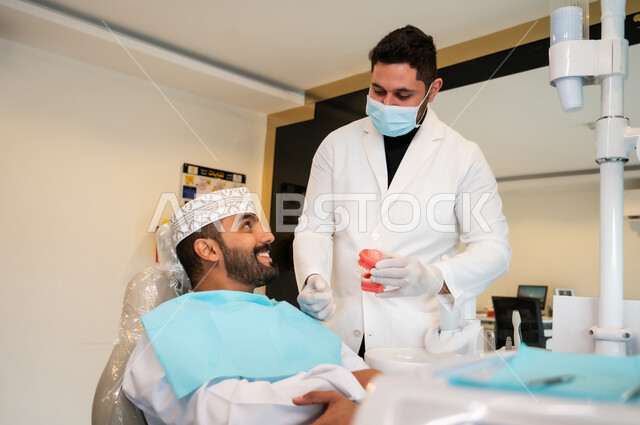 تقديم الرعاية الصحية في المراكز التخصصية، طبيب أسنان يرتدي المعطف والكمامة الطبية مسك بيده نموذج سني، رجل عربي خليجي عماني يجلس في العيادة السنية يتلقى العلاج اللازم للحصول على ابتسامة مثالية بتعابير السرور والاطمئنان، إعطاء الإرشادات والتعليمات اللازمة