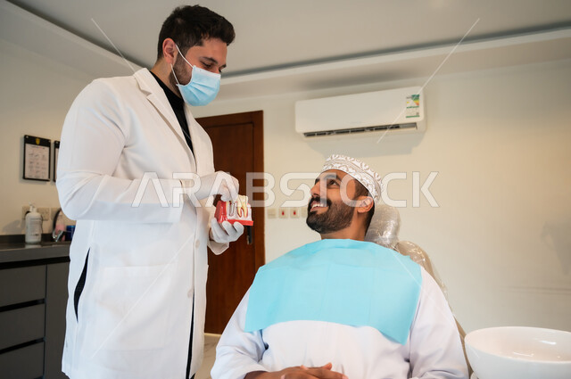 التعليمات والارشادات عن زراعة السن, الاهتمام في الصحة الفموية, النظر إلى الدكتور بايماءات السرور والسعادة, طبيب أسنان يرتدي المعطف والقفازات والكمامة الطبية يعطي نصائح طبية لرجل عربي خليجي عماني يرتدي الزي التقليدي يجلس في العيادة السنية