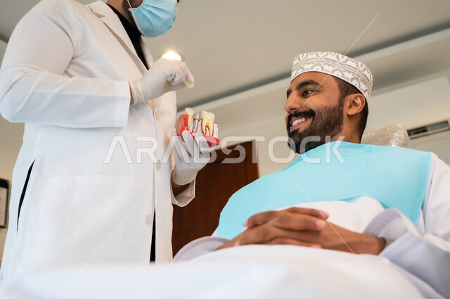 الطرق العلاجية المتبعة للحصول على ابتسامة مثالية، التعليمات والارشادات عن زراعة السن, الاهتمام في الصحة الفموية, رجل عربي خليجي عماني يرتدي الزي التقليدي يجلس في العيادة السنية يتلقى نصائح طبية من طبيب أسنان يرتدي المعطف والقفازات والكمامة