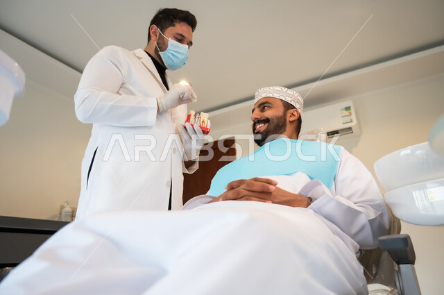 الأساليب العلاجية المتبعة للعناية بالفم، التعليمات والارشادات عن زراعة السن, الاهتمام في الصحة الفموية, طبيب أسنان يرتدي المعطف والقفازات والكمامة الطبية يعطي نصائح طبية لرجل عربي خليجي عماني يرتدي الزي التقليدي يجلس في العيادة السنية