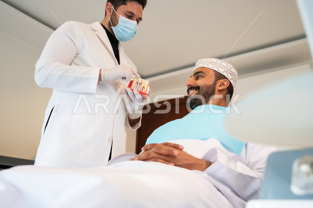 الأساليب العلاجية المتبعة للعناية بالفم، التعليمات والارشادات عن زراعة السن, الاهتمام في الصحة الفموية, طبيب أسنان يرتدي المعطف والقفازات والكمامة الطبية يعطي نصائح طبية لرجل عربي خليجي عماني يرتدي الزي التقليدي يجلس في العيادة السنية