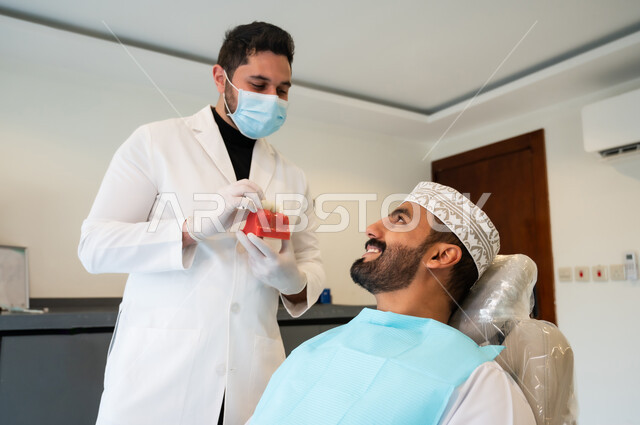 الارشادات والتعليمات عن زراعة السن, الاهتمام في الصحة الفموية, النظر الى الطبيب بايماءات السرور والسعادة, طبيب أسنان يرتدي المعطف و القفازات والكمامة الطبية يعطي نصائح طبية لرجل عربي خليجي عماني يرتدي الزي التقليدي يجلس في العيادة السنية