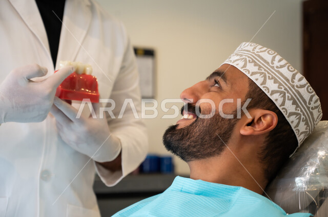 الحصول على ابتسامة مثالية، التعليمات والارشادات عن زراعة السن, الاهتمام في الصحة الفموية, صورة مقربة لرجل عربي خليجي عماني يرتدي الزي التقليدي يجلس في العيادة السنية ينظر إلى طبيب الأسنان بإيماءات السعادة والسرور