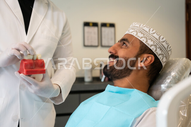 الحصول على ابتسامة مثالية، التعليمات والارشادات عن زراعة السن, الاهتمام في الصحة الفموية, صورة مقربة لرجل عربي خليجي عماني يرتدي الزي التقليدي يجلس في العيادة السنية ينظر إلى طبيب الأسنان بإيماءات السعادة والسرور