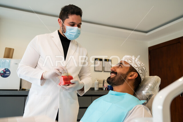 Looking at the doctor with gestures of pleasure and happiness, medical instructions and directions about dental implants, taking care of oral health, a dentist wearing a coat, gloves and a medical mask giving medical advice to an Arab Gulf Omani man wearing traditional clothing sitting in the dental clinic