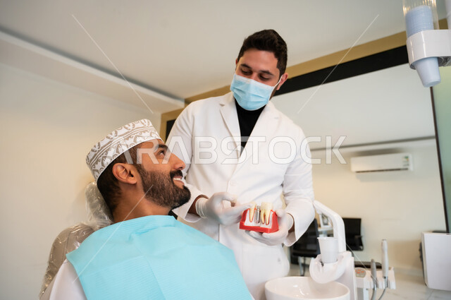 استخدام نموذج صناعي لشرح كيفية زراعة الأسنان، طبيب يرتدي الكمامة والمعطف الطبي يمسك بيده نموذج سني، الاهتمام في الصحة الفموية, مفهوم الطب والرعاية الصحية، صورة مقربه لرجل عربي خليجي عماني يرتدي الزي التقليدي ينظر إلى طبيبه بتعابير الاطمئنان والسرور