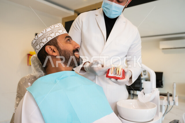 شرح كيفية زراعة الأسنان، طبيب يرتدي الكمامة والمعطف الطبي يمسك بيده نموذج سني، الاهتمام في الصحة الفموية, مفهوم الطب والرعاية الصحية، صورة مقربه لرجل عربي خليجي عماني يرتدي الزي التقليدي ينظر إلى طبيبه بتعابير الاطمئنان والسرور