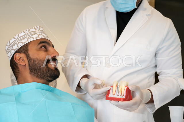 شرح كيفية زراعة الأسنان، طبيب يرتدي الكمامة والمعطف الطبي يمسك بيده نموذج سني، الاهتمام في الصحة الفموية, مفهوم الطب والرعاية الصحية، صورة مقربه لرجل عربي خليجي عماني يرتدي الزي التقليدي ينظر إلى طبيبه بتعابير الاطمئنان والسرور