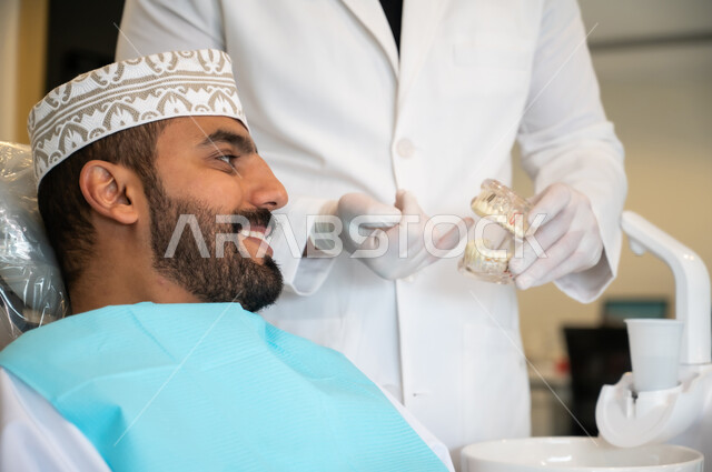 تعابير الاطمئنان والسرور، تقديم نصائح للمحافظة على صحة الفم والأسنان، الاهتمام في الصحة الفموية, مفهوم الطب والرعاية الصحية, طبيب أسنان يرتدي المعطف و القفازات الطبية يمسك بيده نموذج سني، صورة مقربه لرجل عربي خليجي عماني يرتدي الكمه يجلس على كرسي العلاج