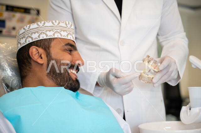 تقديم نصائح للمحافظة على صحة الفم والأسنان، ايماءات الاطمئنان والسرور، الاهتمام في الصحة الفموية, مفهوم الطب والرعاية الصحية, طبيب أسنان يرتدي المعطف و القفازات الطبية يمسك بيده نموذج سني، صورة مقربه لرجل عربي خليجي عماني يرتدي الكمه يجلس على كرسي العلاج