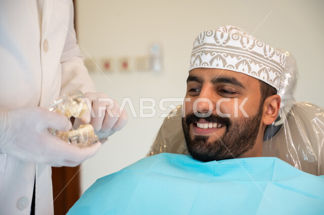 تعابير الاطمئنان والسرور، تقديم نصائح للمحافظة على صحة الفم والأسنان، الاهتمام في الصحة الفموية, مفهوم الطب والرعاية الصحية, طبيب أسنان يرتدي المعطف و القفازات الطبية يمسك بيده نموذج سني، صورة مقربه لرجل عربي خليجي عماني يرتدي الكمه يجلس على كرسي العلاج