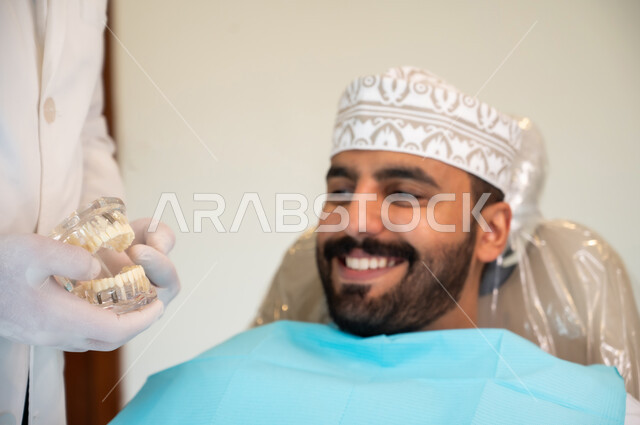 تقديم نصائح للمحافظة على صحة الفم والأسنان، ايماءات الاطمئنان والسرور، الاهتمام في الصحة الفموية, مفهوم الطب والرعاية الصحية, طبيب أسنان يرتدي المعطف و القفازات الطبية يمسك بيده نموذج سني، صورة مقربه لرجل عربي خليجي عماني يرتدي الكمه يجلس على كرسي العلاج
