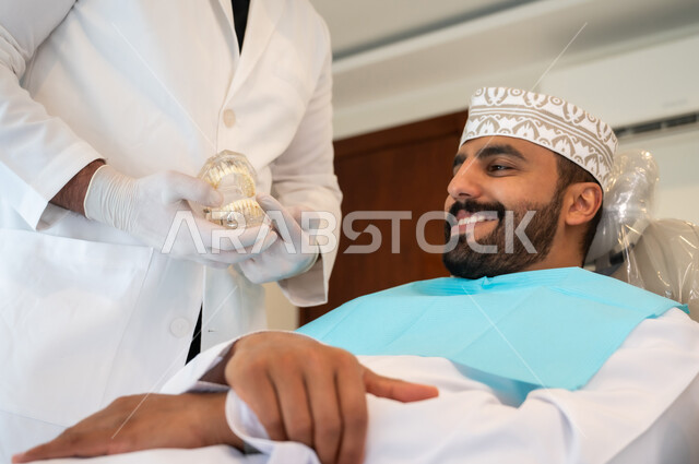 تعابير الاطمئنان والسرور، تقديم نصائح للمحافظة على صحة الفم والأسنان، الاهتمام في الصحة الفموية, مفهوم الطب والرعاية الصحية, طبيب أسنان يرتدي المعطف و القفازات الطبية يمسك بيده نموذج سني، صورة مقربه لرجل عربي خليجي عماني يرتدي الكمه يجلس على كرسي العلاج