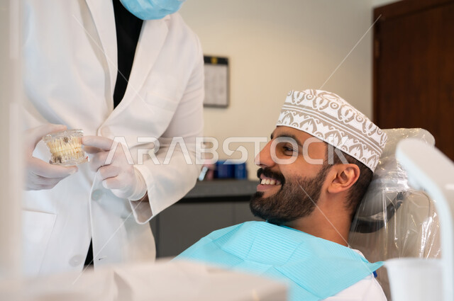 تقديم نصائح للمحافظة على صحة الفم والأسنان، ايماءات الاطمئنان والسرور، الاهتمام في الصحة الفموية, مفهوم الطب والرعاية الصحية, طبيب أسنان يرتدي المعطف و القفازات الطبية يمسك بيده نموذج سني، صورة مقربه لرجل عربي خليجي عماني يرتدي الكمه يجلس على كرسي العلاج
