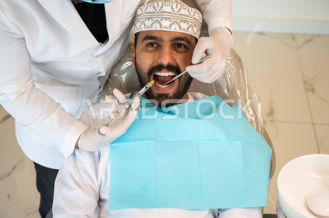 المواد والأدوات اللازمة للتخفيف الشعور بالألم، معالجة النخور والتسوس، صورة ممقربة من الأعلى لرجل عربي خليجي عماني يرتدي الزي التقليدي يجلس على كرسي العلاج في العيادة السنية, طبيب أسنان يرتدي الكمامة والقفازات الطبية يقوم بحقن البنج الموضعي باستخدام الإبرة