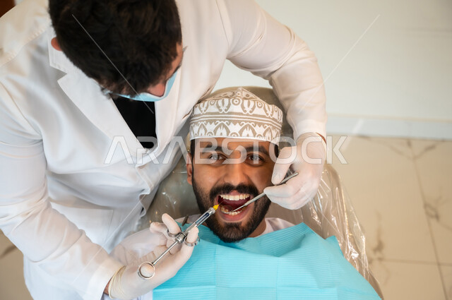المواد والأدوات اللازمة للتخفيف الشعور بالألم، معالجة النخور والتسوس، صورة مقربة من الأعلى لرجل عربي خليجي عماني يرتدي الزي التقليدي يجلس على كرسي العلاج في العيادة السنية, طبيب أسنان يرتدي الكمامة والقفازات الطبية يقوم بحقن البنج الموضعي باستخدام الإبرة