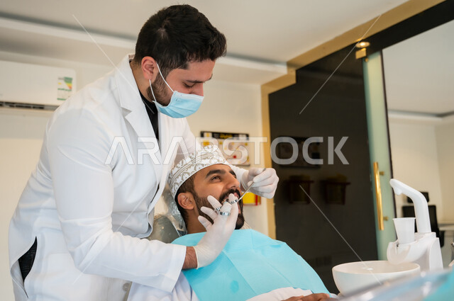 الادوات والمواد اللازمة للتخفيف الشعور بالألم، معالجة النخور والتسوس، رجل عربي خليجي عماني يرتدي الزي التقليدي يجلس على كرسي العلاج في العيادة السنية, طبيب أسنان يرتدي الكمامة والقفازات الطبية يقوم بحقن البنج الموضعي باستخدام الإبرة