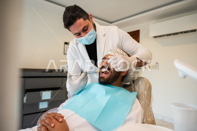 استخدام مادة التخدير للتخفيف الشعور بالألم، معالجة النخور والتسوس، رجل عربي خليجي عماني يرتدي الزي التقليدي يجلس على كرسي العلاج في العيادة السنية, طبيب أسنان يرتدي الكمامة والقفازات الطبية يقوم بحقن البنج الموضعي باستخدام الإبرة