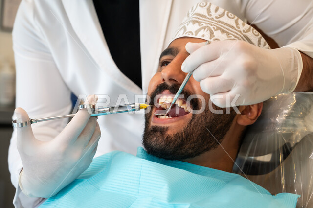 استخدام المعدات والأدوات اللازمة، معالجة النخور والتسوس، مفهوم تخفيف الشعور بالألم , صورة مقربة لرجل عربي خليجي عماني يرتدي الزي التقليدي يجلس في العيادة السنية ويتلقى العلاج اللازم لفمه, طبيب أسنان يرتدي القفازات الطبية يقوم بحقن البنج الموضعي باستخدام الإبرة