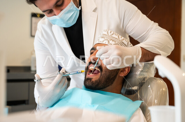 Pain relief, use of necessary equipment and tools, treatment of cavities and decay, close-up of an Omani Gulf Arab man wearing traditional clothing sitting in the dental clinic receiving the necessary treatment for his mouth, a dentist wearing medical gloves injecting local anesthesia using a needle