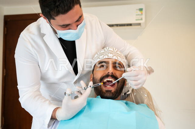 معالجة النخور والتسوس، مفهوم تخفيف الشعور بالألم , صورة مقربة لرجل عربي خليجي عماني يرتدي الزي التقليدي يجلس في العيادة السنية ويتلقى العلاج اللازم لفمه, طبيب أسنان يرتدي الكمامة والقفازات الطبية يقوم بحقن البنج الموضعي باستخدام الإبرة