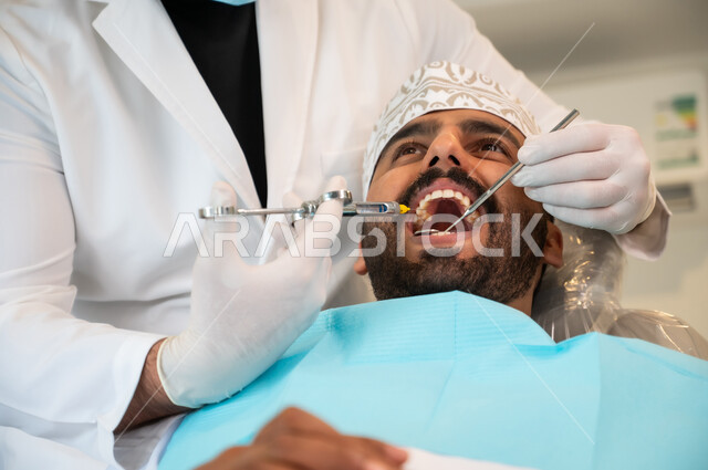 استخدام المعدات والأدوات اللازمة، معالجة النخور والتسوس، مفهوم تخفيف الشعور بالألم , صورة مقربة لرجل عربي خليجي عماني يرتدي الزي التقليدي يجلس في العيادة السنية ويتلقى العلاج اللازم لفمه, طبيب أسنان يرتدي القفازات الطبية يقوم بحقن البنج الموضعي باستخدام الإبرة