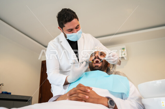 استخدام مادة التخدير للتخفيف الشعور بالألم، معالجة النخور والتسوس، رجل عربي خليجي عماني يرتدي الزي التقليدي يجلس على كرسي العلاج في العيادة السنية, طبيب أسنان يرتدي الكمامة والقفازات الطبية يقوم بحقن البنج الموضعي باستخدام الإبرة