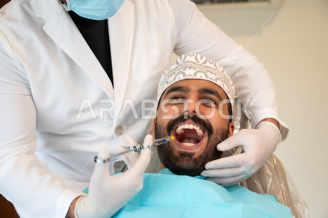معالجة النخور والتسوس، مفهوم تخفيف الشعور بالألم , صورة مقربة لرجل عربي خليجي عماني يرتدي الزي التقليدي يجلس في العيادة السنية ويتلقى العلاج اللازم لفمه, طبيب أسنان يرتدي الكمامة والقفازات الطبية يقوم بحقن البنج الموضعي باستخدام الإبرة