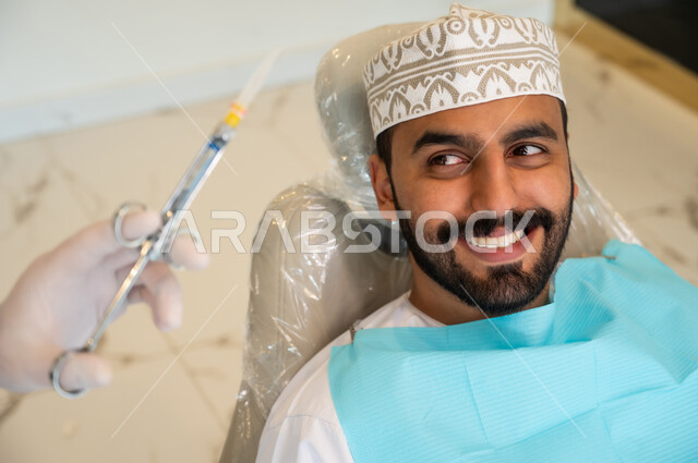 الاستعداد لحقن البنج الموضعي لتخفيف الشعور بالألم ,تقديم الرعاية الصحية في المراكز التخصصية، صورة مقربة لرجل عربي خليجي عماني يرتدي الزي التقليدي يجلس في العيادة السنية بإيماءات السرور, يد طبيب أسنان يرتدي القفازات الطبية يمسك بيده إبرة المخدر،معالجة النخور والتسوس