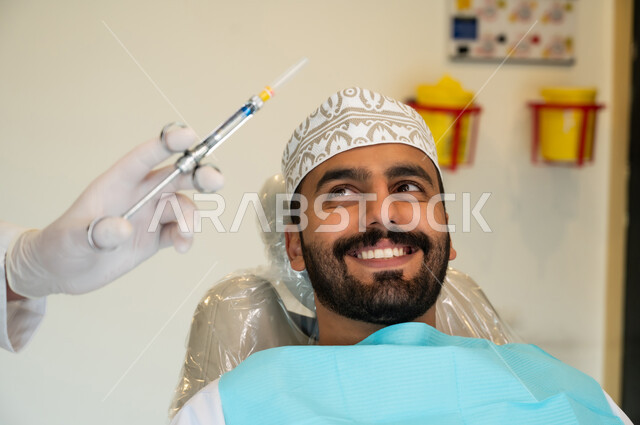 معالجة النخور والتسوس، الاستعداد لحقن البنج الموضعي لتخفيف الشعور بالألم ,تقديم الرعاية الصحية في المراكز التخصصية، صورة مقربة لرجل عربي خليجي عماني يرتدي الزي التقليدي يجلس في العيادة السنية بإيماءات السرور, يد طبيب أسنان يرتدي القفازات الطبية يمسك بيده إبرة المخدر