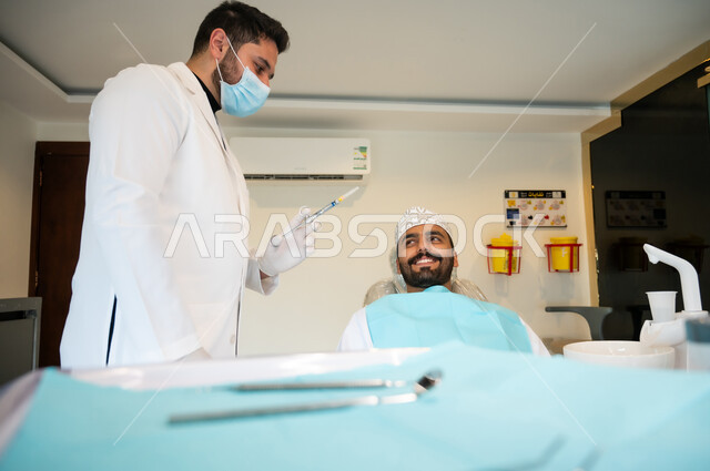 استخدام المعدات والأدوات اللازمة لمعالجة النخور والتسوس، الاستعداد لحقن البنج الموضعي لتخفيف الشعور بالألم، رجل عربي خليجي عماني يرتدي الزي التقليدي يجلس في العيادة السنية بإيماءات السرور, يد طبيب أسنان يرتدي المعطف والقفازات الطبية يمسك بيده إبرة المخدر