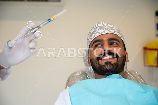 معالجة النخور والتسوس، الاستعداد لحقن البنج الموضعي لتخفيف الشعور بالألم ,تقديم الرعاية الصحية في المراكز التخصصية، صورة مقربة لرجل عربي خليجي عماني يرتدي الزي التقليدي يجلس في العيادة السنية بإيماءات السرور, يد طبيب أسنان يرتدي القفازات الطبية يمسك بيده إبرة المخدر