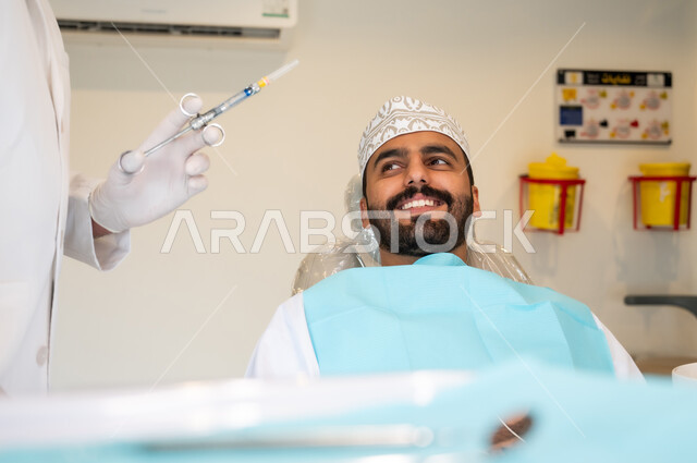 الاستعداد لحقن البنج الموضعي لتخفيف الشعور بالألم ,تقديم الرعاية الصحية في المراكز التخصصية، صورة مقربة لرجل عربي خليجي عماني يرتدي الزي التقليدي يجلس في العيادة السنية بإيماءات السرور, يد طبيب أسنان يرتدي القفازات الطبية يمسك بيده إبرة المخدر،معالجة النخور والتسوس