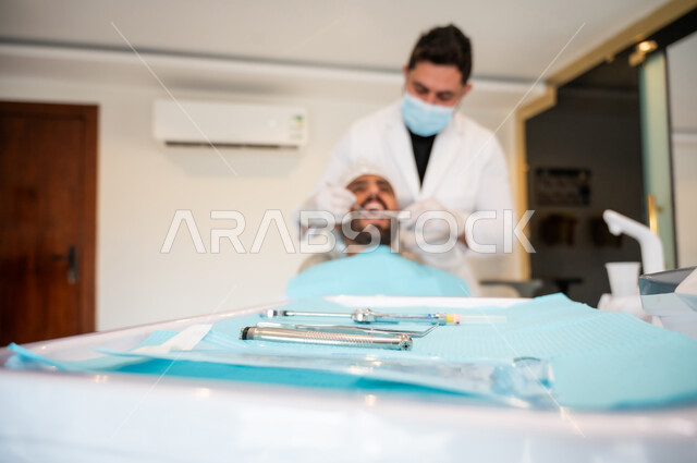 زيارة العيادات الصحية، صورة مقربة للمعدات والأدوات اللازمة لجراحة الفم, طبيب يقوم بفحص أسنان المريض تشخيص المرض، إجراء الفحوصات الدورية للعناية بالصحة الفموية, معالجة التسوس وتركيب الحشوات، شاب عربي خليجي عماني يرتدي الزي التقليدي يجلس على كرسي العلاج