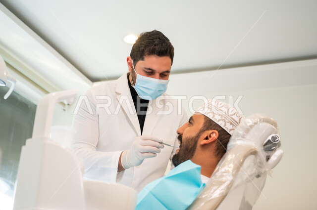 استخدام المعدات اللازمة لجراحة الفم, معالجة النخور وتركيب الحشوات، الزيارة الدورية للاطمئنان على الصحة الفموية، طبيب يرتدي المعطف والكمامة الطبية يقوم بفحص أسنان المريض و تشخيص المرض، صورة من الجانب لشاب عربي خليجي عماني يرتدي الكمة يجلس في العيادة السنية