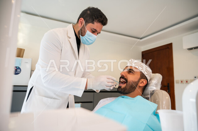 إجراء الفحوصات للعناية بالصحة الفموية، استعمال المعدات اللازمة لجراحة الفم, معالجة النخور وتركيب الحشوات، طبيب يرتدي القفازات والكمامة الطبية يقوم بفحص أسنان المريض و تشخيص المرض، شاب عربي خليجي عماني يرتدي الكمة يجلس في العيادة السنية