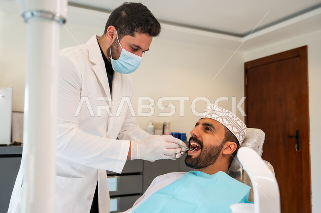 استعمال المعدات اللازمة لجراحة الفم, معالجة النخور وتركيب الحشوات، طبيب يرتدي القفازات والكمامة الطبية يقوم بفحص أسنان المريض و تشخيص المرض، صورة مقربة لشاب عربي خليجي عماني يرتدي الكمة يجلس في العيادة السنية يقوم بإجراء الفحوصات للعناية بالصحة الفموية