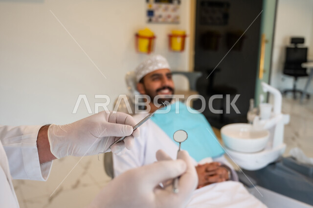 زيارة العيادات الصحية، صورة مقربة ليدين طبيب يمسك المعدات والأدوات اللازمة لفحص أسنان المريض وتشخيص المرض، إجراء الفحوصات الدورية للعناية بالصحة الفموية, معالجة التسوس وتركيب الحشوات، شاب عربي خليجي عماني يرتدي الزي التقليدي يجلس على كرسي العلاج