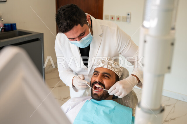 معالجة التسوس وتركيب الحشوات، إجراء الفحوصات الدورية للعناية بالصحة الفموية، زيارة العيادات الصحية، طبيب يرتدي الكمامة والقفازات الطبية يقوم بفحص أسنان المريض وتشخيص المرض, صورة من الأعلى لشاب عربي خليجي عماني يرتدي الزي التقليدي يجلس على كرسي العلاج