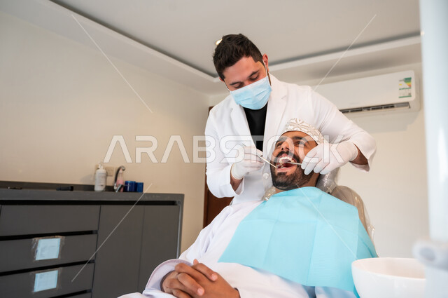 معالجة التسوس وتركيب الحشوات، إجراء الفحوصات الدورية للعناية بالصحة الفموية، طبيب يرتدي الكمامة والقفازات الطبية يقوم بفحص أسنان المريض وتشخيص المرض, شاب عربي خليجي عماني يرتدي الزي التقليدي يجلس على كرسي العلاج في العيادة السنية