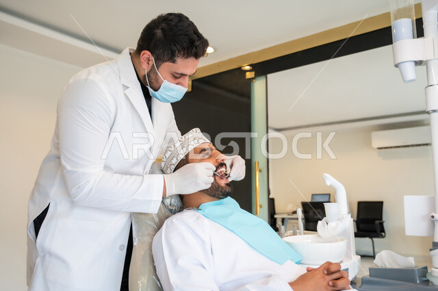 Visiting the dental clinic for regular checkups, fillings, and tooth decay treatment, taking care of oral health, a doctor wearing a mask and medical gloves examining the patient's teeth and diagnosing the disease, a young Arab Gulf Omani man wearing traditional clothing sitting on a treatment chair