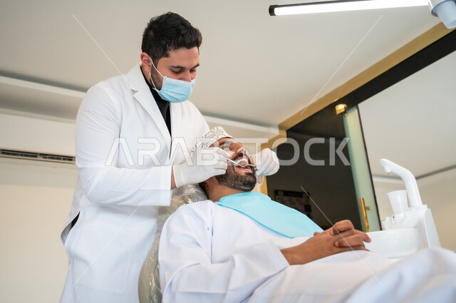 إجراء الفحوصات الدورية في العيادة السنية، تركيب الحشوات ومعالجة التسوس، العناية بالصحة الفموية، طبيب يرتدي الكمامة والقفازات الطبية يقوم بفحص أسنان المريض وتشخيص المرض, شاب عربي خليجي عماني يرتدي الزي التقليدي يجلس على كرسي العلاج