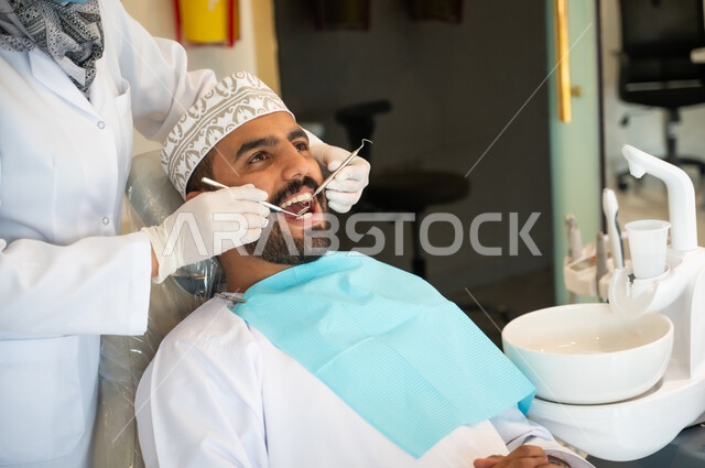 جراحة الفم والفكين، المعدات والادوات اللازمة لإجراء الفحوصات، معالجة النخر والتسوس, شاب عربي خليجي عماني يرتدي الزي التقليدي يجلس في العيادة السنية ويفتح فمه لتشخيص المرض, طبيبة عربية خليجيه عمانية ترتدي القفازات الطبية تقوم بفحص أسنان المريض