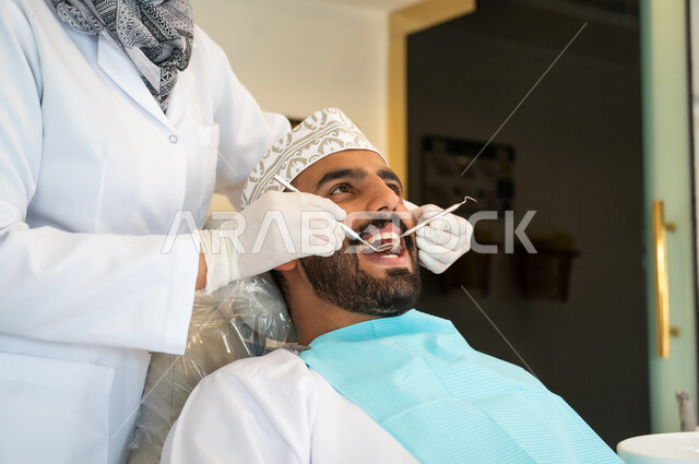 المعدات والادوات اللازمة لإجراء الفحوصات، معالجة النخر والتسوس, صورة مقربة لشاب عربي خليجي عماني يرتدي الزي التقليدي يجلس في العيادة السنية ويفتح فمه لتشخيص المرض, طبيبة عربية خليجيه عمانية ترتدي القفازات الطبية تقوم بفحص أسنان المريض
